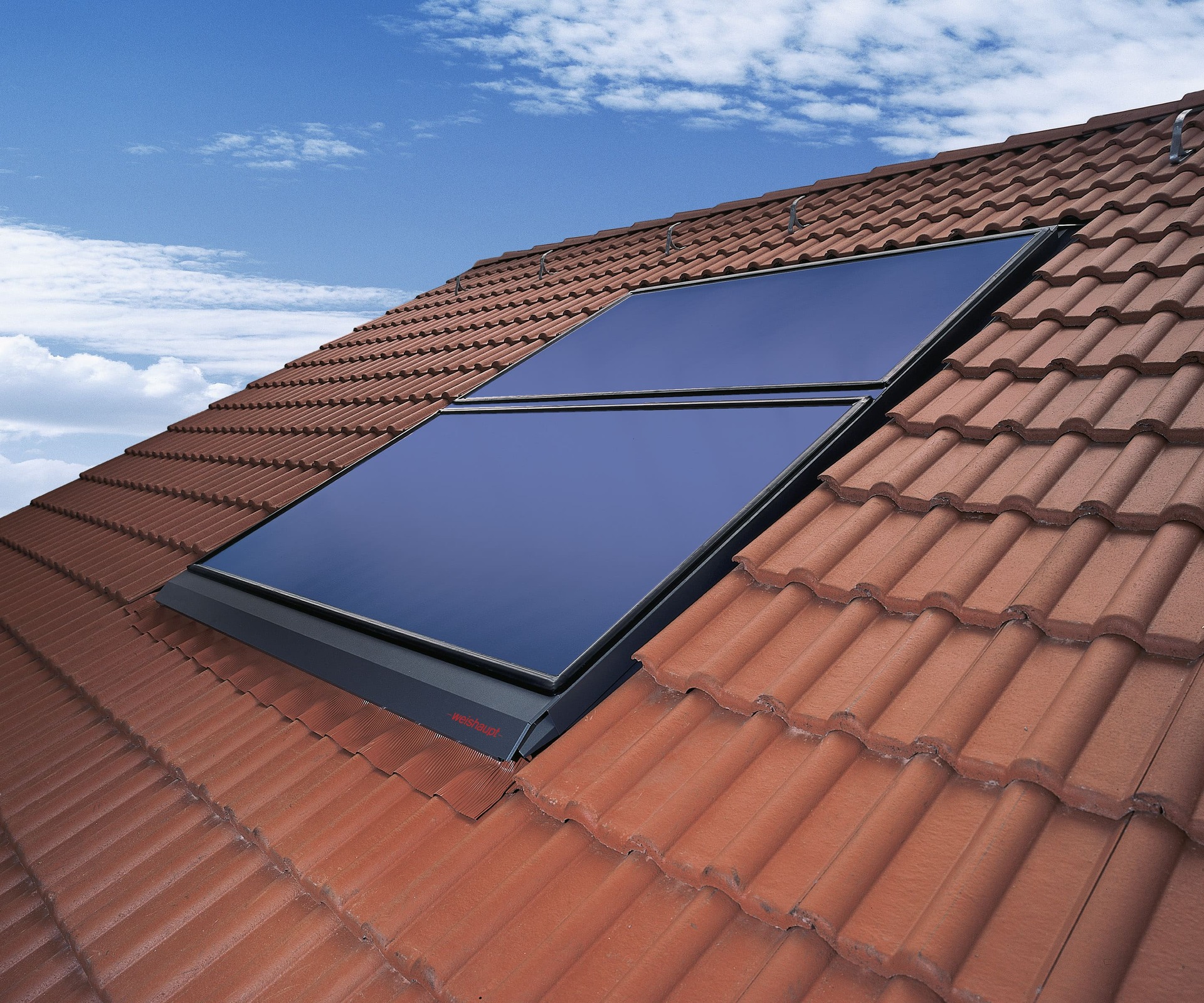 Solartechnik Ein Foto, das zwei große, rechteckige, dunkelblaue Solarthermie-Kollektoren zeigt, die in eine rote Ziegeldachfläche integriert sind. Die Kollektoren sind flächenbündig in das Dach eingelassen und von einem schwarzen Rahmen umgeben. Die Ziegel des Daches sind in einem Fischgrätmuster verlegt. Im Hintergrund ist ein blauer Himmel mit weißen Wolken zu sehen. Das Foto ist aus einer leicht erhöhten Perspektive aufgenommen.